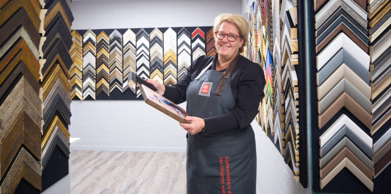 Natascha Heijnen met een ingelijst werk bij de stalenwand met honderden lijstprofielen in het atelier